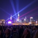 Diverse crowd in city square, bright lights, global landmarks.